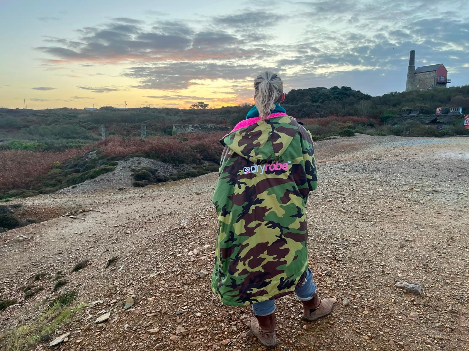Candace wrapped in a changing robe on a Cornish clifftop path at sunrise, looking out over gorse and an old engine house