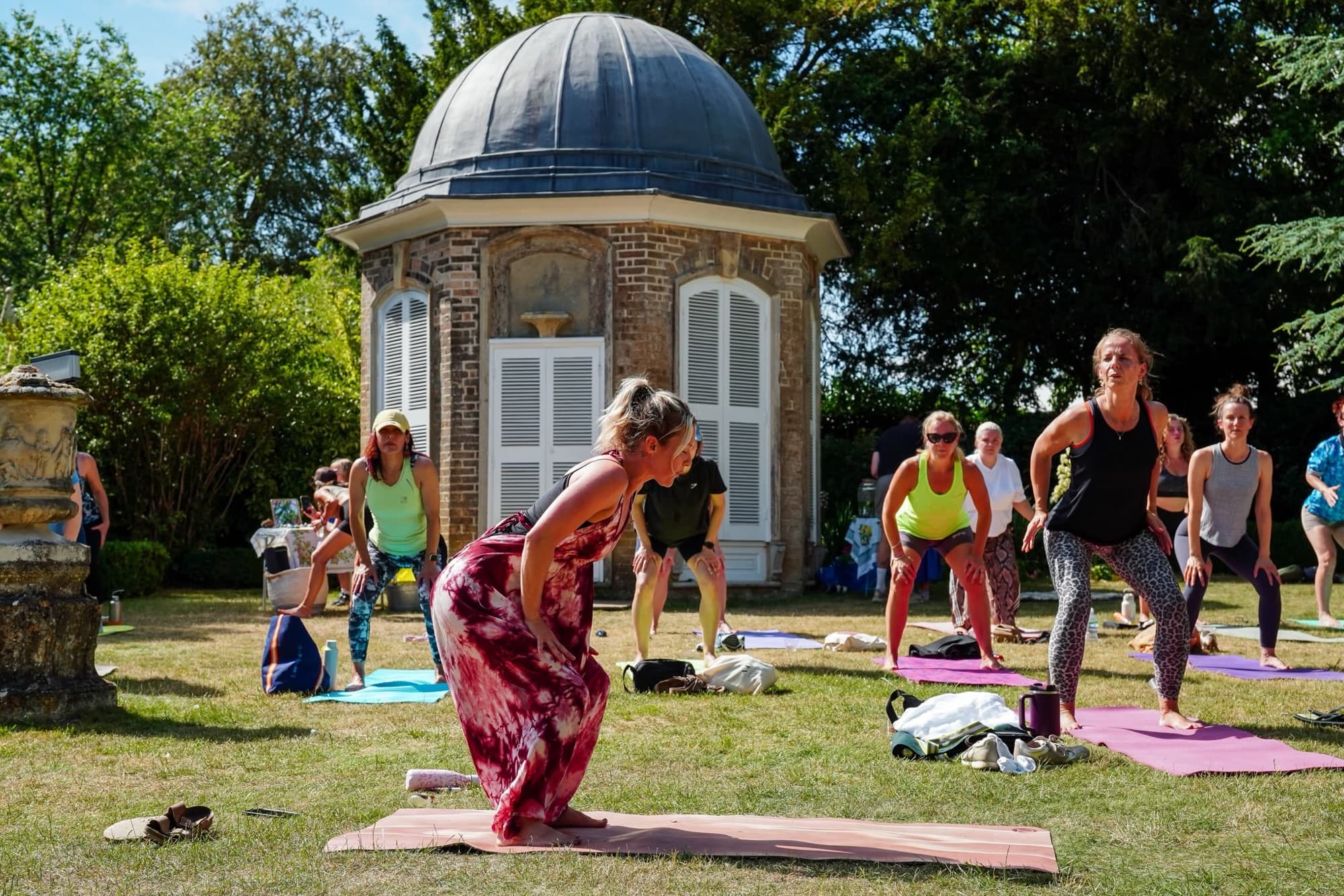 Candace Redden leading an outdoor yoga class on the lawn in front of a domed garden pavilion, students folding forward on their mats in summer sunshine