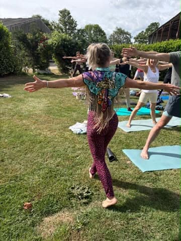 Candace guiding a standing balance with arms open wide while students mirror the shape behind her on a sunlit lawn